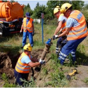 Na zdjęciu widać trzech pracowników w pomarańczowych kamizelkach odblaskowych i żółtych kaskach, którzy wykonują prace ziemne związane z infrastrukturą wodociągową. Dwóch z nich podnosi metalową rurę, a trzeci pomaga w jej wydobyciu z wykopu. W tle znajduje się pracownik w niebieskiej czapce oraz pojazd asenizacyjny z pomarańczowym zbiornikiem. Teren wokół wykopu jest porośnięty trawą i roślinnością.